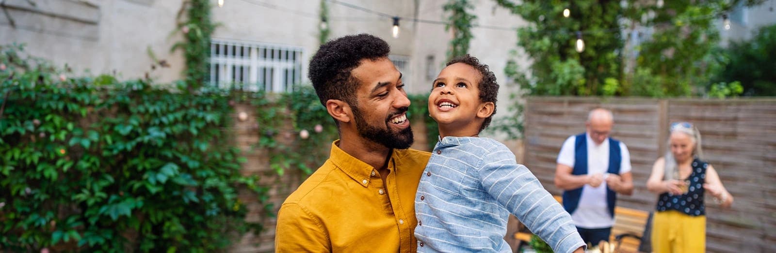 A father holding his young son in a backyard, with the boy's grandparents nearby